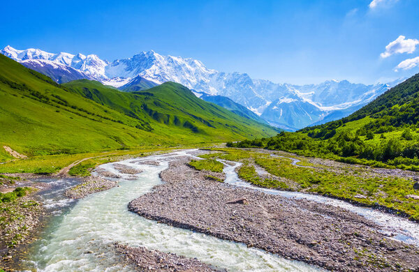 From the river shore, covered with stones, opens view on fantastic glacier and steep rocky mountains with green meadows, which are covered with snow. Happy lifestyle. Beautiful spring background.