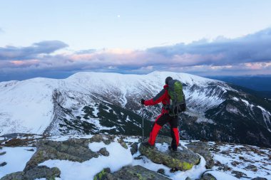 Büyük bir sırt çantası ve seyahat sopa ile bir spor muhabiri Çimenlerin arasında bir kaya üzerinde duruyor ve inanılmaz karla kaplı dağ manzara ve mavi gökyüzü görünüyor.