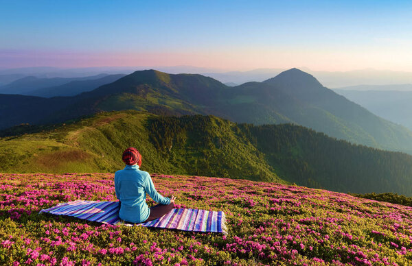 On the lawn among the rhododendron flowers the yoga girl is sitting in the pose on the colorful rug. Meditation. The landscape with the mountains in the fog. Nice summer scenery.