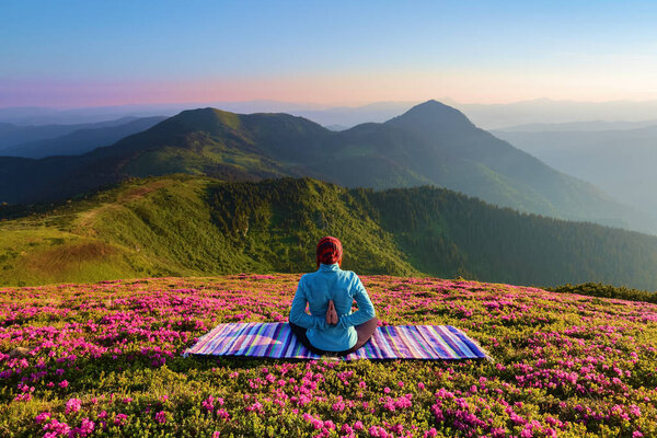 Colorful mat. The yoga girl in the lotus pose. The lawn with the rhododendron flowers. High mountains. Magical forest. Meditation. Relax. Summer scenery. Warm morning sun rays.