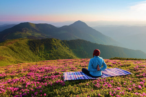 Colorful carpet. The yoga girl in the lotus pose. The lawn with the rhododendron flowers. High mountains. Magical forest. Meditation. Relax. Summer scenery. Warm morning sun rays.