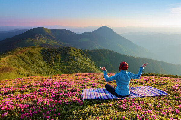 On the lawn among the rhododendron flowers the yoga girl is sitting in the pose on the colorful rug. Meditation. The landscape with the mountains in the fog. Nice summer scenery.