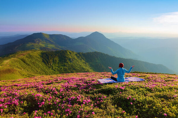 Colorful karemat. The yoga girl in the lotus pose. The lawn with the rhododendron flowers. High mountains. Magical forest. Meditation. Relax. Summer scenery. Warm morning sun rays.