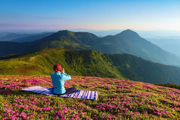 Colorful karemat. The yoga girl in the lotus pose. The lawn with the rhododendron flowers. High mountains. Magical forest. Meditation. Relax. Summer scenery. Warm morning sun rays.