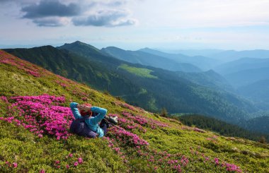 Manzara yüksek dağlarla. Gökyüzü bulutlu. Orman yolu. Orman gülleri çimenlerin üzerine kız yatıyor. Eko turizm. Büyüleyici yaz gün.