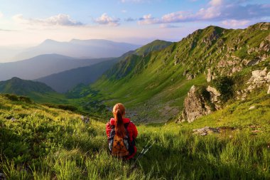 O görünümle yüksek rocky Dağları. Genç bir kız yeşil çimen çiçekler arasında oturur. Sur ışınları. Güzel bulutlar gökyüzünde yavaş yavaş hareket ediyor. Yaz sahne.