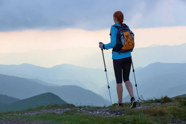 The tourist girl with back sack and tracking sticks is staying on the lawn. Beautiful cloudy sky. Sun rays fall down on the mountains. Summer scene. Eco touristic laisure time.