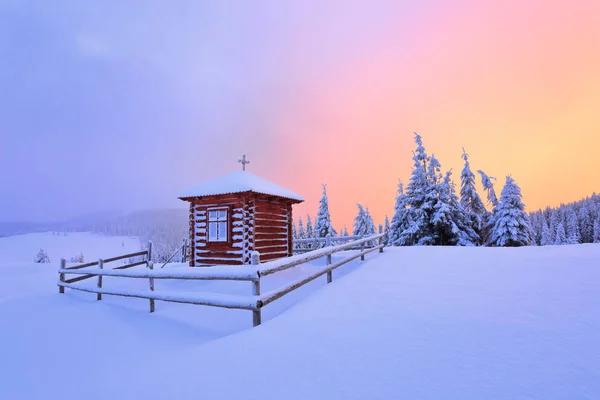 素晴らしい風景が日没とバラの空の素敵な雪に覆われた木の芝生から開かれます 芝生の上は 高山に積もった雪で覆われた木造の教会 — ストック写真