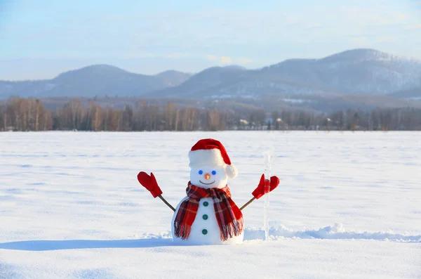 Happy snowman with ice pikestaff is standing on the snow lawn. Field in snow. Mountains on the background. Cold winter day.