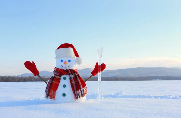 The snowman in plaid scarf, red hat, gloves and icicle in the hand. Nice landscape with the mountains on the background. Field covered with snow. Cold winter day.
