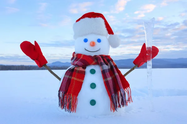 The snowman in plaid scarf, red hat, gloves. Amazing sunrise enlighten the sky. Nice landscape with the mountains on the background. Field covered with snow.