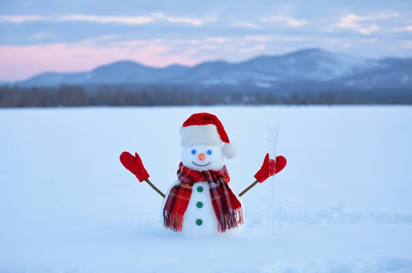 The snowman in plaid scarf, red hat, gloves. Amazing sunrise enlighten the sky. Nice landscape with the mountains on the background. Field covered with snow.