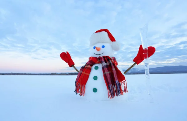The blue eyed smiling snowman in red hat, gloves and plaid scarf holds the icicle in hand. Joyful cold winter morning. Field in snow.