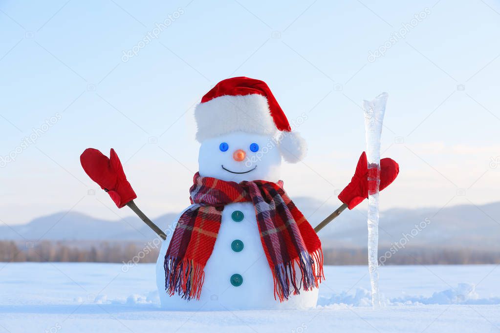 Happy snowman with ice pikestaff is standing on the snow lawn. Field in snow. Mountains on the background. Cold winter day.