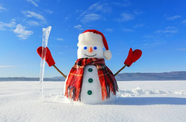 The snowman in plaid scarf, red hat, gloves and icicle in the hand. Nice landscape with the mountains on the background. Field covered with snow. Cold winter day.