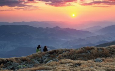 Bir çift aşık kayaların üzerinde oturan ve sahne ile bulutlar gökyüzü günbatımı, yüksek dağların izliyor. Turuncu ve kırmızı renkleri ile ufuk yanar.   