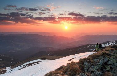 Sahne şaşırtıcı günbatımı, gökyüzü bulutlu ile kırmızı ve turuncu renkleri ile aydınlatılır. Bir kaç içinde aşk kayaların üzerinde oturuyor. Karda çim.   