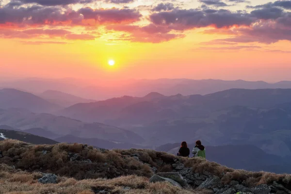A couple in love is sitting on the rocks and watching the scenery of the sunset, high mountains, sky with clouds. The horizon lights up with orange and red colors.