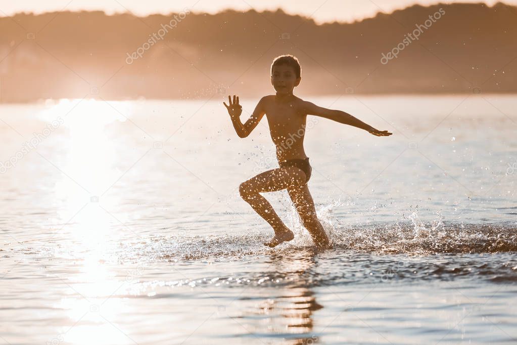 On the nice sunny summer day the small boy is playing, running, jumping at the lake. Happy moments of childhood. Reflection of the silhouette on the water. Beautiful place Svityaz lake, Ukraine.