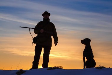 Bir silah ve onun Alman Shorthaired Pointer köpek cins arkadaşı ile şapka bir avcı. Şafağın arka planında güzel siluetler. İnanılmaz kış sabahı.