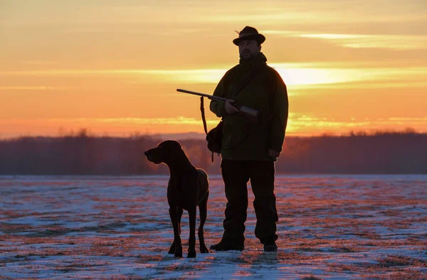 A hunter in a hat with a gun and a German shorthair pointer dog breed stands on a meadow covered with snow. Majestic winter scenery. Amazing sunrise. — Stock Photo, Image