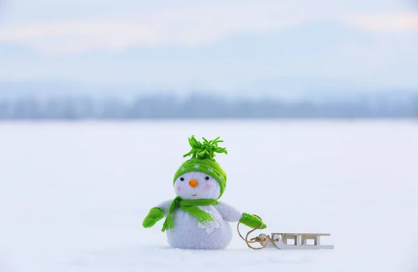 Snowman in green hat, gloves and scarf pulls the sledge. Christmas concept with snowy background. Beautiful winter day. Happy new year. — Stock Photo, Image