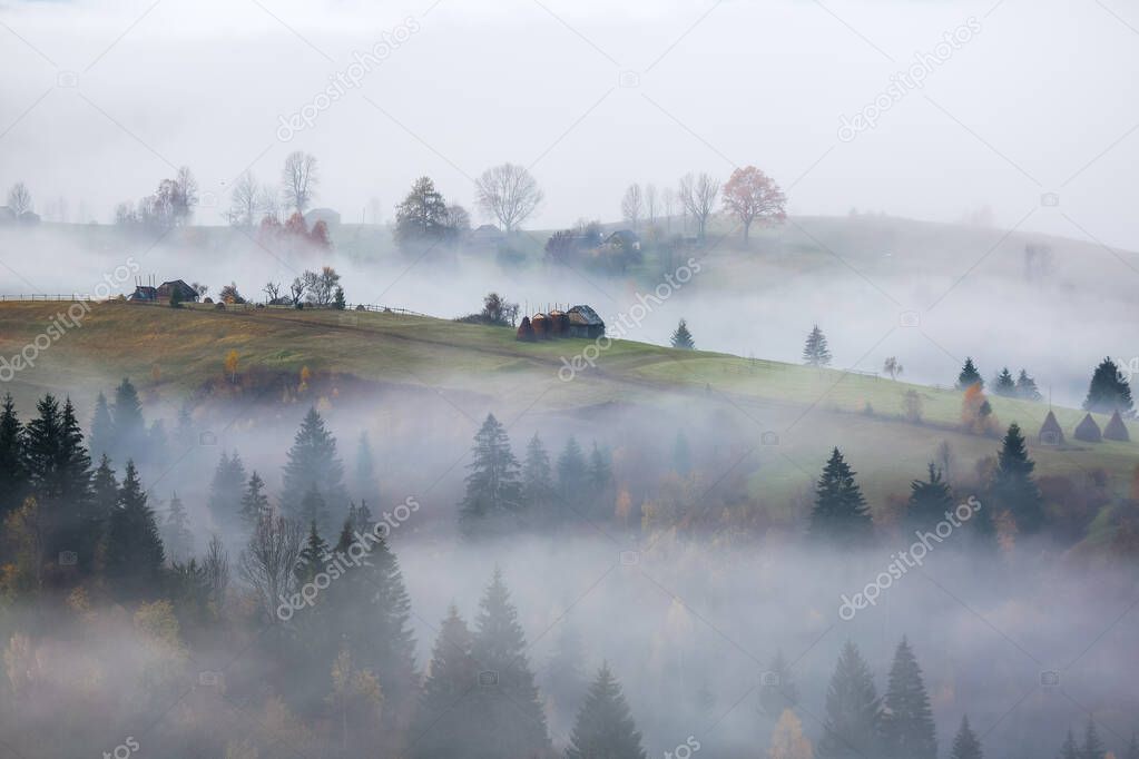 Autumn scenery with the fruit trees, haystacks and house. Countryside, fields, forests in the morning fog. Landscape with the mountains.