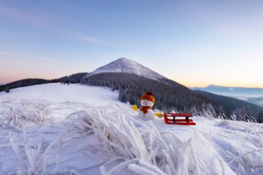 Şapkalı kardan adam, eldivenler ve atkılar kızağı çeker. Güzel bir kış günü. Mutlu yıllar. Çim karla kaplı. Yüksek dağlı manzara. Mutlu yıllar..