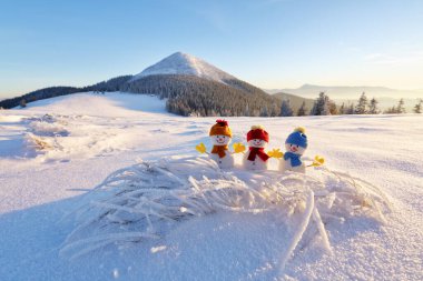 Karlı çimlerde şapkalı ve atkılı üç küçük kardan adam. Güzel kış günbatımı arka planı. Yüksek dağlı manzara. Mutlu Noeller ve mutlu yıllar..