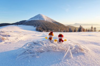 Kar tarlasında kırmızı şapkalı ve eşarplı iki küçük kardan adam. Güzel kış günbatımı arka planı. Yüksek dağlı manzara. Mutlu Noeller ve mutlu yıllar..