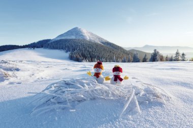 Kar tarlasında kırmızı şapkalı ve eşarplı iki küçük kardan adam. Güzel kış günbatımı arka planı. Yüksek dağlı manzara. Mutlu Noeller ve mutlu yıllar..