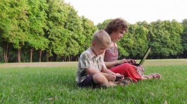 Anne ve oğlu gadget'lar ile parkta çimlerde oturuyor. Annem bir dizüstü bilgisayar, bir çocuk bir gadget ile çalışıyor..