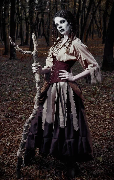 Halloween theme: ugly scary voodoo witch with stick. Portrait of the evil sorceress in dark forest. Zombie woman (undead)