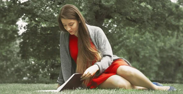 Beautiful woman reading book in nature. Young attractive student in red dress studying and reading a book. (Relax, rest, education concept,  recreation).