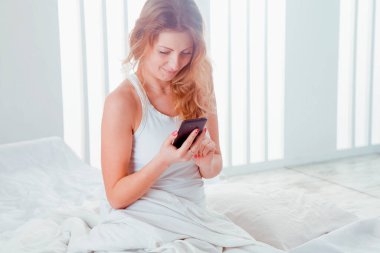 Günaydın iyi haberle başlıyor. Yatakta akıllı telefondan haber izleyen mutlu genç bir kadının portresi..