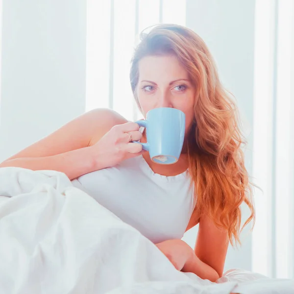 Good morning and good mood starts with a delicious morning coffee.  Young beautiful woman sitting on bed holding a cup of coffee.
