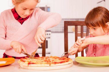 İki küçük bayan arkadaş pizzayı çatal ve bıçakla kesiyor. Mutlu çocuklar yemek yerken eğleniyor. Yatay resim.