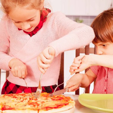İki küçük bayan arkadaş pizzayı çatal ve bıçakla kesiyor. Mutlu çocuklar yemek yerken eğleniyor..