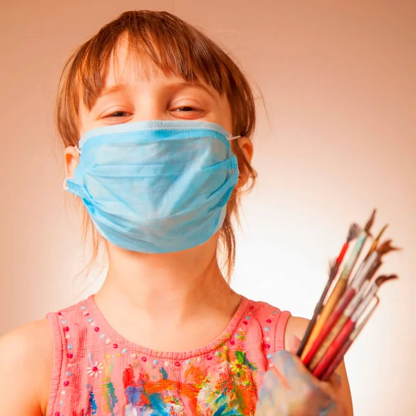 Online painting lessons during quarantine. Close up portrait of cute little child girl with protect medical face mask learn to painting with finger. Virus, health and remotely education concept.