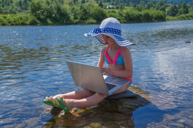 Genç, güzel, serbest çalışan bir kadının nehir taşına oturmuş dizüstü bilgisayar kullanırken çekilmiş komik bir fotoğrafı.. 