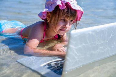 Her zaman ve her yerde öğrenmek. Küçük tatlı bir kızın komik portresi tropikal denizde dizüstü bilgisayarla uzaktan öğreniyor..