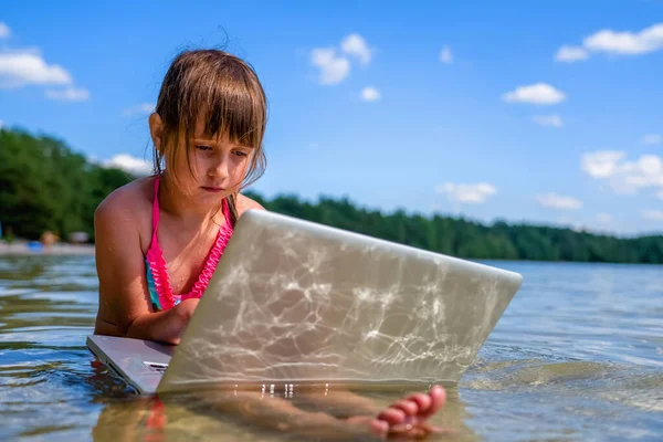 Her zaman ve her yerde çalışmak. Küçük tatlı kızın komik portresi dışarıda tropikal denizin sularında dizüstü bilgisayarla çalışıyor..