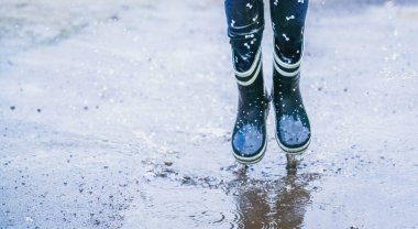 Sonbahar yağmurlu hava. Yağmurdan sonra yağmur botlarıyla su birikintilerine atlayan bir çocuk. Seçici odaklanma. Yatay resim.