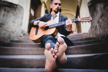 Beyaz adam basamaklarda oturup gitar çalıyor ve şarkı söylüyor. Ayaklara seçici odaklanma.