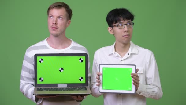 Два молодых мультиэтнических бизнесмена показывают на камеру ноутбук и цифровой планшет — стоковое видео