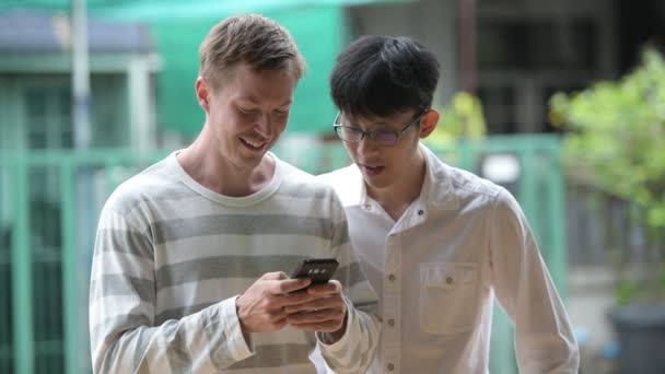 Deux hommes d'affaires multiethniques heureux souriant tout en utilisant le téléphone ensemble dans les rues à l'extérieur — Video