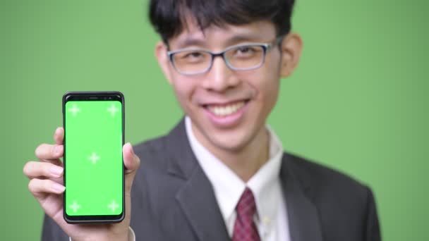 Junge glückliche asiatische Geschäftsmann lächelt, während zeigt Telefon in die Kamera — Stockvideo