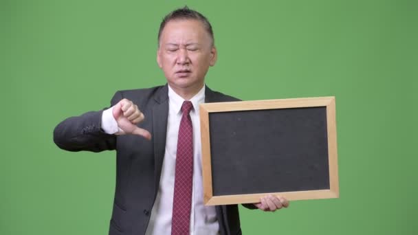 Mature Japanese businessman holding blackboard against green background — Stock Video