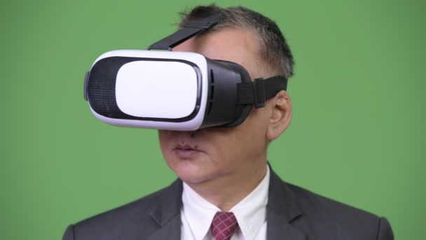 Hombre de negocios japonés maduro usando auriculares de realidad virtual contra fondo verde — Vídeo de stock