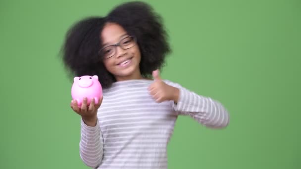 Jovem bonito menina africana com cabelo afro segurando porquinho banco e dando polegares para cima — Vídeo de Stock